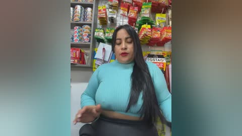 Video of a Latina woman with long black hair, wearing a turquoise ribbed crop top and black pants, standing in a convenience store with shelves filled with snacks and drinks in the background.