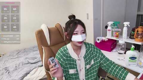 A video of an Asian woman with dark hair tied in a bun, wearing a green Chanel jacket and a white surgical mask, holding a thermometer, seated on a brown chair in a messy room with cleaning supplies and a gray bed.
