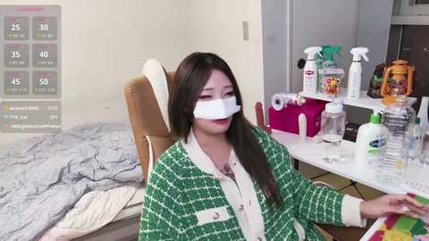 Video of a woman wearing a white face mask, green plaid cardigan, and beige shirt, sitting at a cluttered desk with cleaning supplies, surrounded by a messy room.
