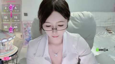 A video of an Asian woman with fair skin, wearing glasses, a white blouse, and a butterfly necklace, sitting on a bed, in a bedroom setting.