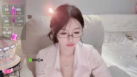 Video of an East Asian woman with fair skin, wearing glasses, a white blouse, and a necklace, sitting on a beige couch. Background includes a table with a vase and a white bed.