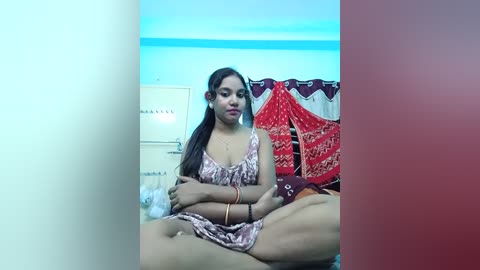 A video of a young woman with medium skin tone and long dark hair, wearing a patterned dress, sitting cross-legged on a bed in a brightly lit room with colorful wall hangings.