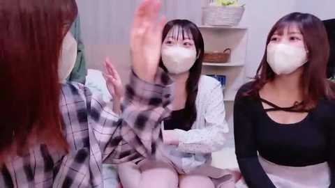 Video of three East Asian women in face masks, wearing casual clothes, seated indoors. One raises her arm, and all have a neutral expression.