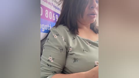 Video of a plus-sized woman with medium-length brown hair, wearing a light green floral-patterned blouse, standing in a room with a white and blue background.