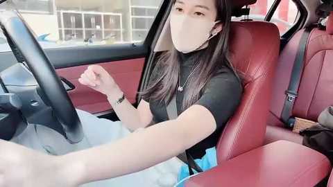A video of a young woman with long black hair, wearing a black top, mask, and headphones, sitting in a red leather car seat, clearing snow from the windshield.