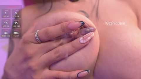 Video of a woman with fair skin, large breasts, and long, manicured nails in a soft, pastel-colored room.