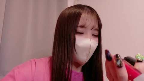 Video of a young Asian woman with straight black hair and pale skin, wearing a pink shirt and a white face mask, applying nail polish with dark nails in a dimly lit room.