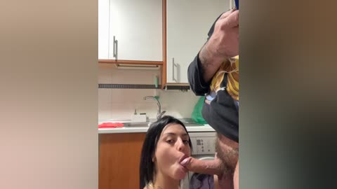 Video of a woman with dark hair, wearing a yellow top, performing oral sex on a man in a kitchen with white cabinets and a sink.