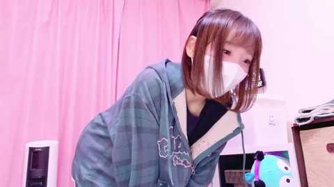 Video of an Asian woman with straight, shoulder-length brown hair, wearing a green plaid jacket, white face mask, and pink curtains in a brightly lit room.