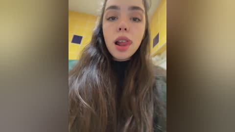 Video of a young woman with long, wavy brown hair, wearing a black top, and smiling slightly. Background features yellow walls with blue and white patterned decorations.