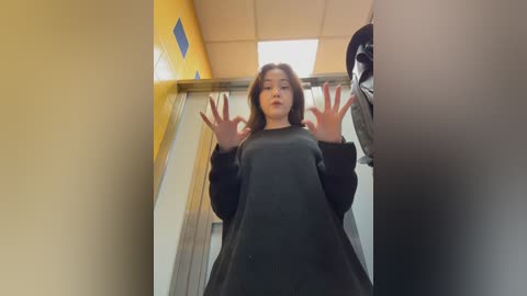 Video of a young woman with shoulder-length brown hair, wearing a black oversized sweater, standing in a narrow, brightly-lit hallway with yellow walls, making a \"peace\" sign with her hands.