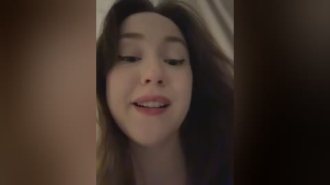 Video of a young, light-skinned woman with long brown hair, wearing minimal makeup, smiling gently, with a blurred background.