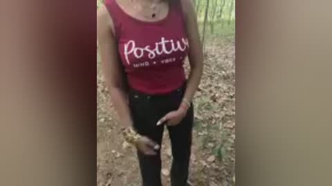 Video of a woman in a red tank top with \"Positive Vibes Only\" text, black ripped jeans, standing outdoors on a dirt path surrounded by greenery.