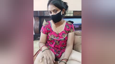 Video of a woman with medium skin tone and dark hair, wearing a pink floral dress, black face mask, and red bracelets, sitting on a beige couch, in a domestic setting.