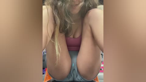 Video of a young woman with light brown hair, wearing a pink tank top and blue shorts, sitting with knees bent and legs spread, creating a \"V\" shape in the center, set against a blurred, indoor background.