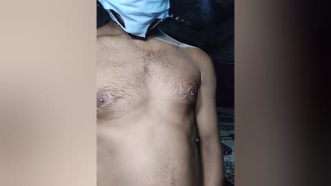 Video of a shirtless, light-skinned, male torso with visible nipples, wearing a blue face mask, standing in a dimly lit room with dark walls and patterned floor.