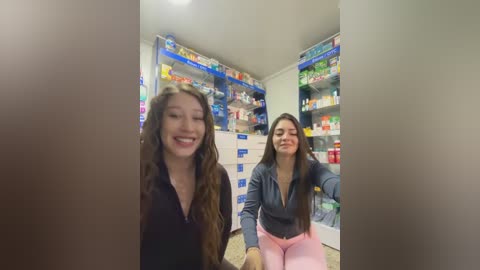 Video of two smiling young women, one with long brown hair in a black top, the other with long black hair in a gray top, standing in a well-stocked pharmacy aisle.