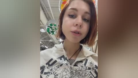 Video of a young woman with straight, shoulder-length brown hair, wearing a white hoodie with a black and gray graphic design. She stands indoors, in front of a ceiling-mounted circular light fixture and a visible ventilation system.