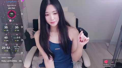 Video of a young East Asian woman with long black hair and fair skin, wearing a blue sleeveless dress, seated in a modern, minimalistic room with white furniture and a mirror.