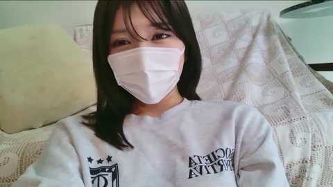 Video of a young Asian woman with straight black hair, wearing a white face mask and a white hoodie with black text, lying on a bed with a white, patterned blanket.