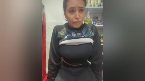 Video of a Latina woman with medium skin tone, dark hair tied back, wearing a black crop top with a blue dragon design, and black pants, in a cluttered kitchen with cabinets and red fridge.