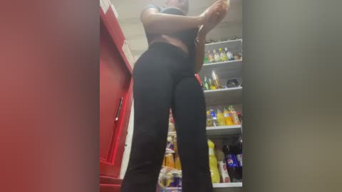 Video of a woman in a red shirt and black leggings standing in a cluttered kitchen with shelves filled with various household items.