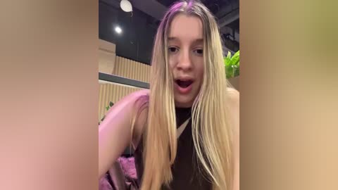 Video of a young Caucasian woman with long, straight blonde hair, wearing a sleeveless black top, posing indoors with a playful expression, surrounded by blurred objects and ambient lighting.