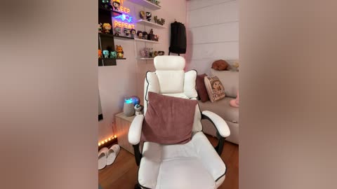 Video of a cozy gaming room with a white gaming chair, purple blanket, plush toys, and neon signs; a woman's feet are visible on the floor.