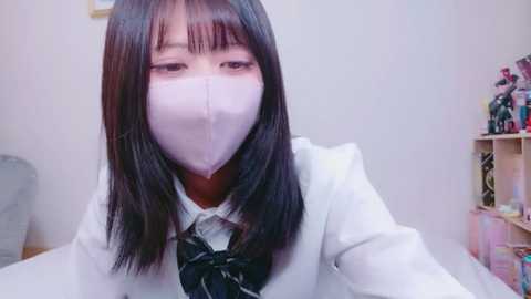 Video of an Asian woman with straight black hair, wearing a white mask, a white blazer, and a black tie, indoors, against a plain white wall, with shelves filled with toys in the background.