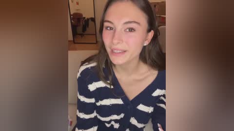 Video of a young woman with fair skin, brown hair, and a warm smile, wearing a navy blue, white-striped cardigan, sitting indoors.