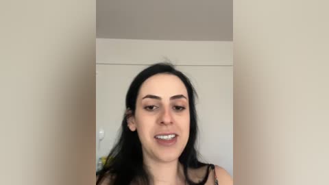 A video of a young woman with long black hair and fair skin, wearing a black lace top, smiling, against a plain white wall and door background.