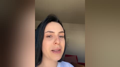 Video of a young woman with fair skin and dark hair, smiling slightly, indoors against a plain beige wall.