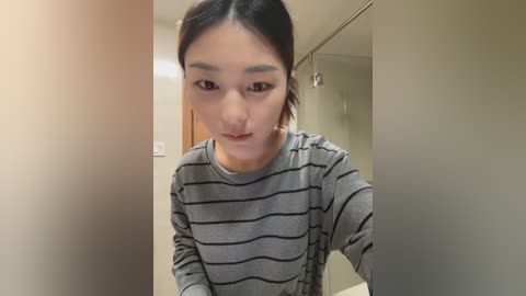 A video of a young East Asian woman with short black hair, wearing a grey and black striped sweater, standing in a minimalist, beige-walled bathroom with a shower door visible in the background.