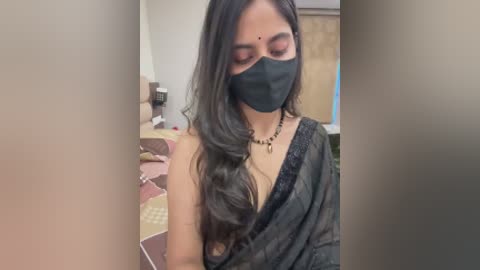 Video of a South Asian woman with long black hair, wearing a black mask, black blouse, and a black necklace, standing in a room with a brown couch and wooden furniture.