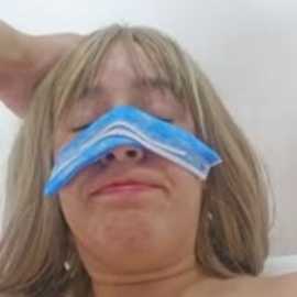 A video of a young woman with long blonde hair, wearing a blue surgical mask that covers her eyes and nose, resting her arm behind her head.