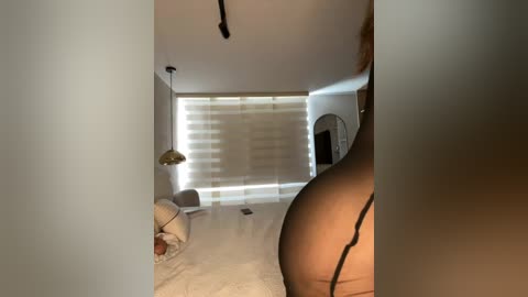 Video of a dimly-lit, minimalist bedroom with white walls, a beige bedspread, a partially visible person with a large, round buttocks, and a window with horizontal blinds.