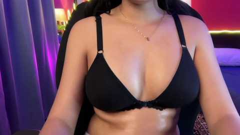 Video of a light-skinned woman with black hair, wearing a black lace bra, sitting in a black chair with purple and pink background.