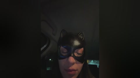 Video of a woman wearing a black Batman mask, with closed eyes, in a dimly lit room with dark shadows.