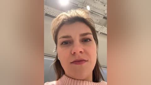 A close-up video of a young woman with fair skin, straight brown hair, and light makeup, wearing a pink turtleneck sweater, taken indoors with a white ceiling and exposed ductwork in the background.