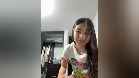 Video of a young Asian woman with long black hair, wearing a colorful graphic t-shirt, standing in a modern, minimalist bathroom with a white sink and dark wood cabinets.