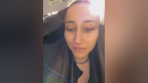 Video of a young woman with fair skin and dark hair, wearing a black choker necklace and a black jacket, captured in a dimly lit room.