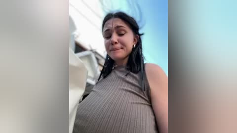 A low-angle video of a Latina woman with long black hair, wearing a ribbed tank top that accentuates her large breasts. She has a neutral expression, and the background features a white building with a blue sky.