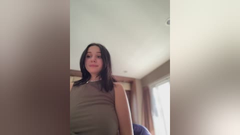 A video of a young woman with long, dark hair, wearing a sleeveless beige top, standing indoors near a window. The background features a modern, light-colored room with wooden accents.