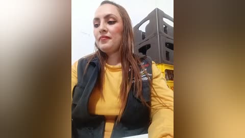 Video of a young woman with light skin, long brown hair, and a yellow top, standing next to a grey speaker. The background is a white wall with a lens flare effect.