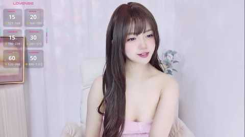 Video of an East Asian woman with long brown hair, light skin, and pink lips, wearing a strapless pink dress, standing in a softly lit room with a calendar and decorative items in the background.