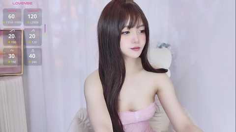 A video of an Asian woman with long black hair, wearing a pink strapless dress, sitting on a white chair in a room with a temperature gauge and a plush toy.