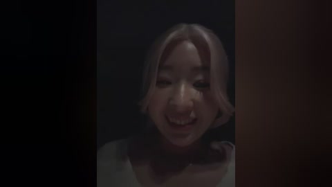 Video of a smiling East Asian woman with shoulder-length light brown hair, wearing a white top, set against a dark background.