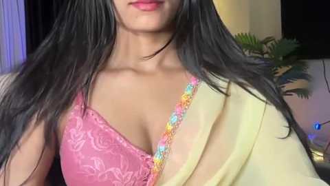 Video of a South Asian woman with long black hair, wearing a pink lace bra and yellow shawl, against a blurred indoor background with green plants.
