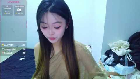 A video of a young Asian woman with long black hair, wearing a beige top, sitting on a bed with scattered clothes. The room is dimly lit, with a digital clock and video recording indicator visible.
