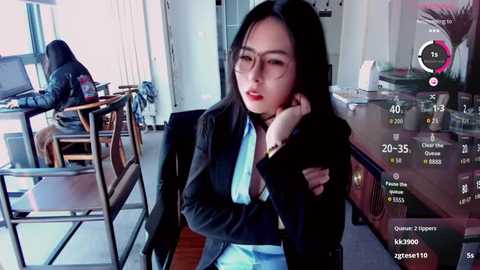 Video of a young Asian woman with long black hair, wearing glasses and a black suit, sitting at a table in a modern office.